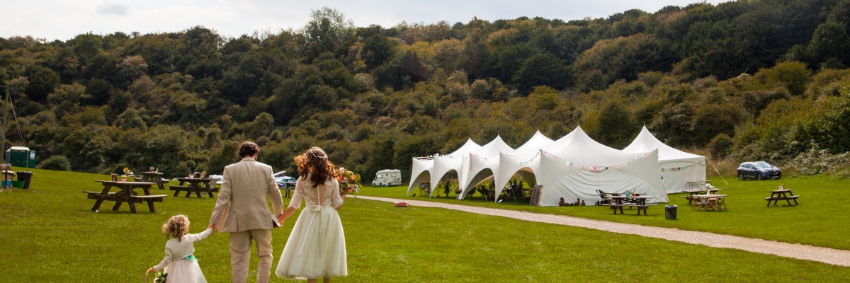 A bridal couple with a young child walking towards a row of marquees in a field set up for a wedding