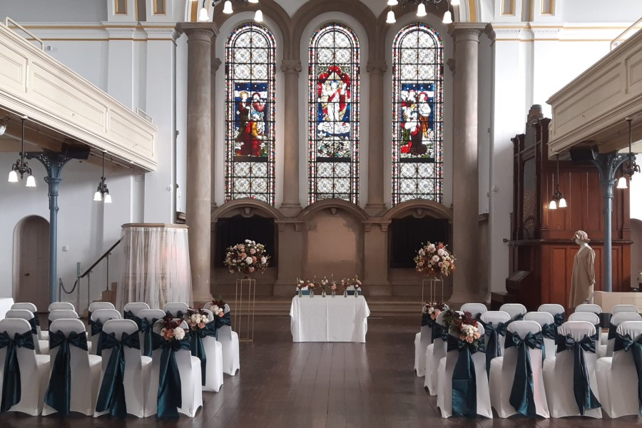 Inside view of Royal Victoria Chapel set up for a wedding ceremony with rows of chairs and a top table in front of a stained glass window