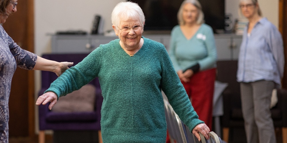 Older lady in a green jumper being assisted during a balance class