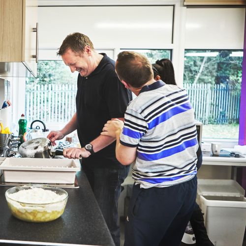 two men in kitchen area smiling, one of the men is washing up