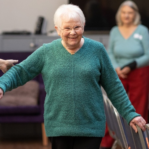 Older lady in a green jumper being assisted during a balance class