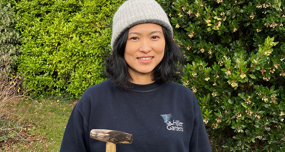 A lady wearing a woolly hat and a blue Sir Harold Hillier Gardens top stood outside and holding gardening tools. 