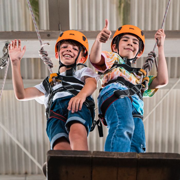 Two children wearing safety helmets and harnesses are standing on a high platform, with their arms waving.