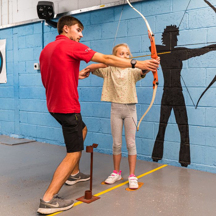 An instructor in a red shirt helps a student with archery indoors. A silhouette of an archer is painted on the blue wall behind them.