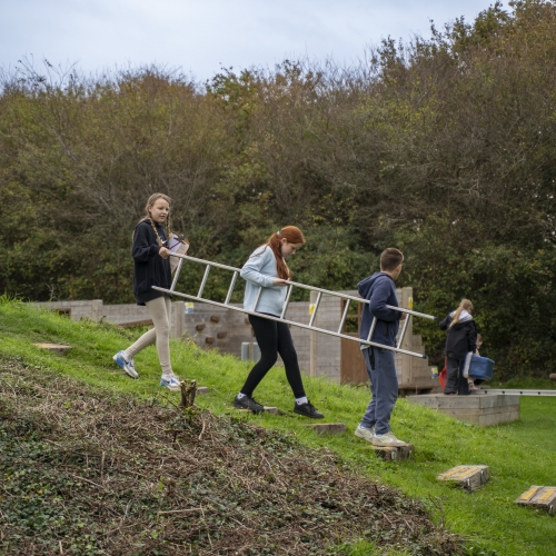Three children working together to carry a long ladder down a slope