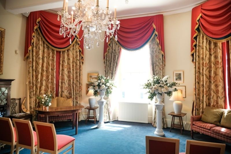 Elegant wedding ceremony room at Serle's House with floral draped curtains, crystal chandelier, guest seating and white flower arrangements.