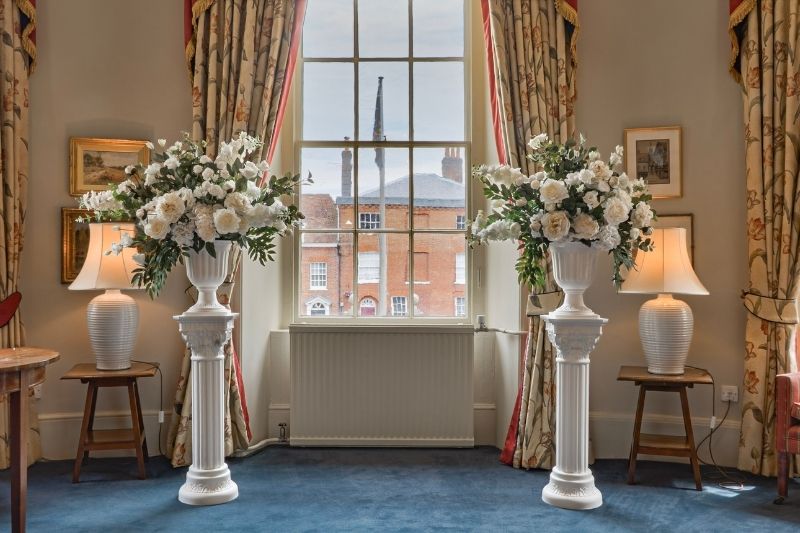 Two large white floral arrangements on pedestal stands positioned in front of a tall window in the Serle's House ceremony room.