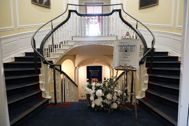 Grand staircase at Serle's House decorated with floral arrangements and a wedding welcome sign beside sweeping curved bannisters.