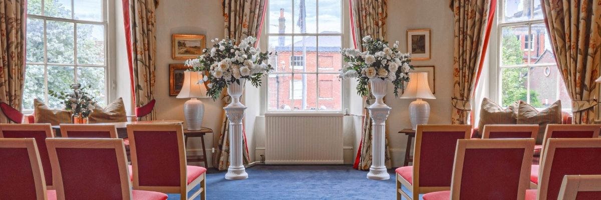 Elegant ceremony room at Serle's House featuring rows of red chairs, a white aisle runner, floral arrangements, ornate curtains and a crystal chandelier.