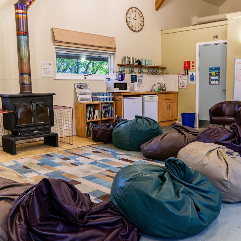 Common area at Minstead Study Centre with several beanbag chairs arranged on a patterned rug, a wood‑burning stove on the left, and a small kitchenette with a sink, fridge, shelves and mugs along the back wall.